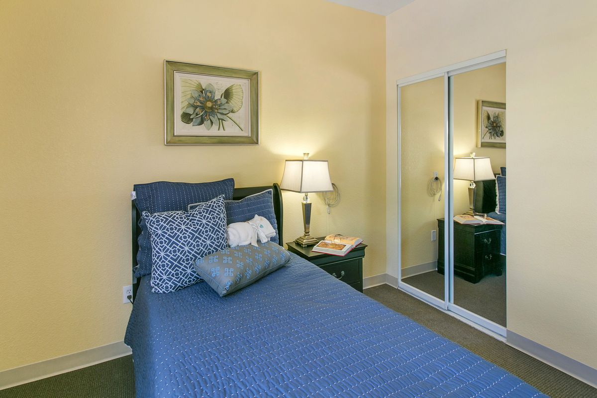 A cozy bedroom with a single bed dressed in blue bedding, accompanied by a nightstand and decorative lamps. A mirrored closet reflects the warm tones of the room.