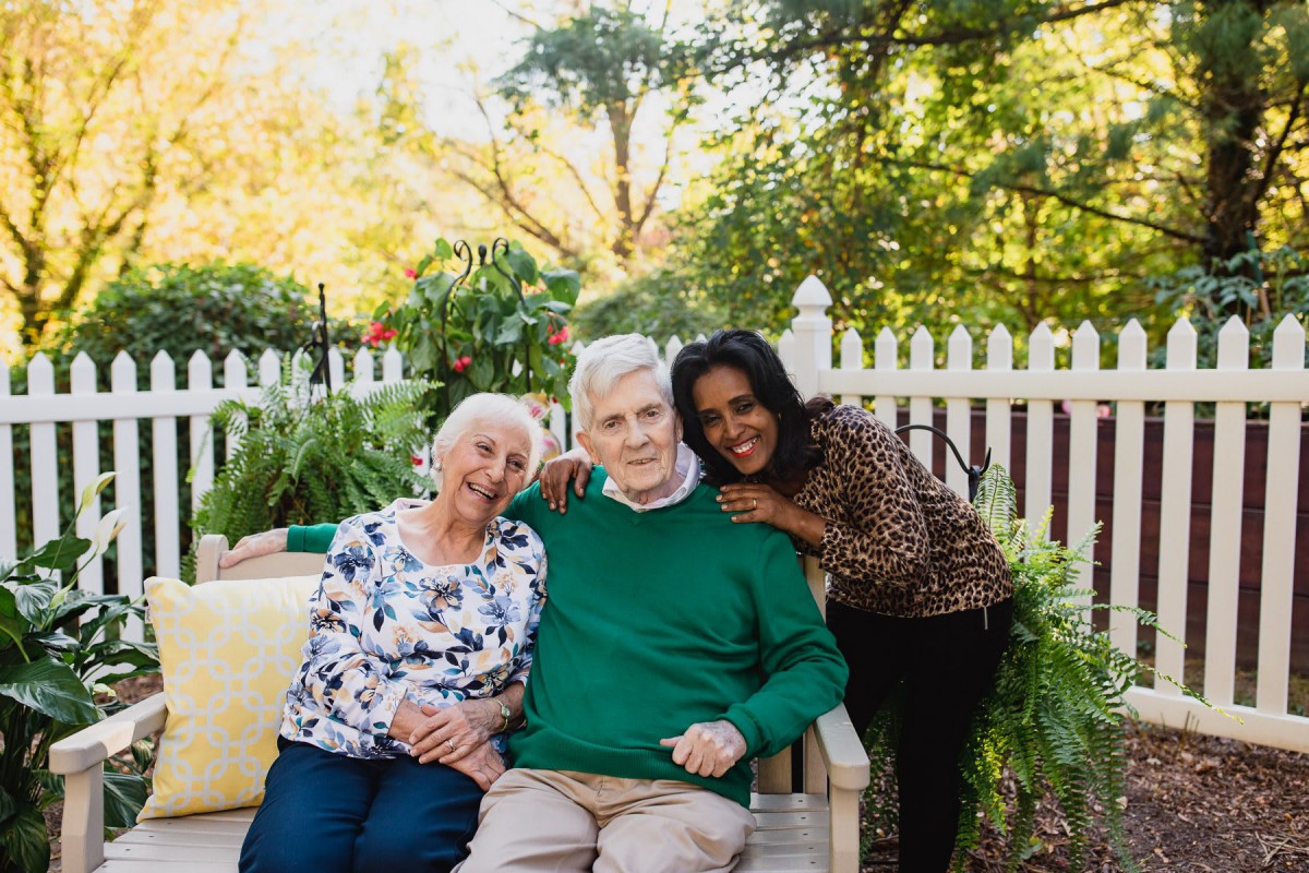 Three people sitting on a bench in a garden, smiling and enjoying each other's company.