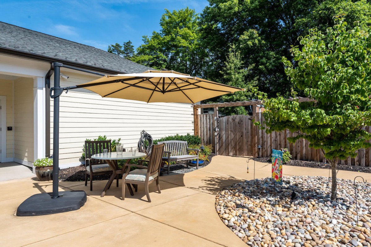 A sunny backyard patio with outdoor furniture and an umbrella, surrounded by a garden and fence.