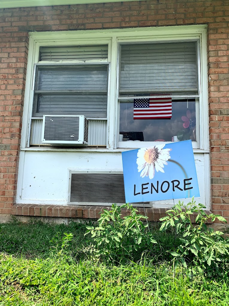 A window with an air conditioner and an American flag, below which is a sign featuring a flower and the name 'Lenore', set against a brick wall and surrounded by grass.