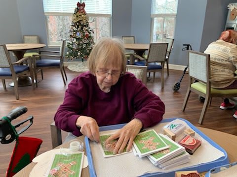 An elderly woman engages in crafting activities at a table with holiday cards, with a Christmas tree in the background.