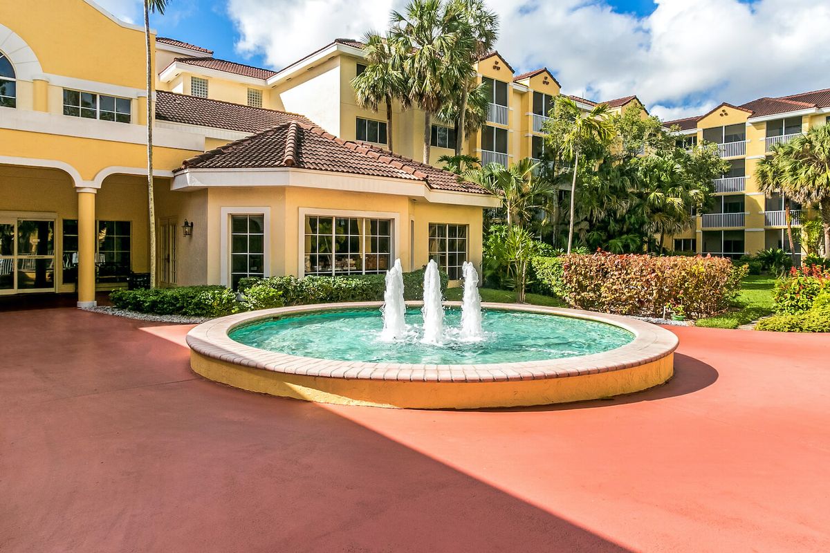 A sunny day view of a hotel entrance with a decorative fountain and palm trees.