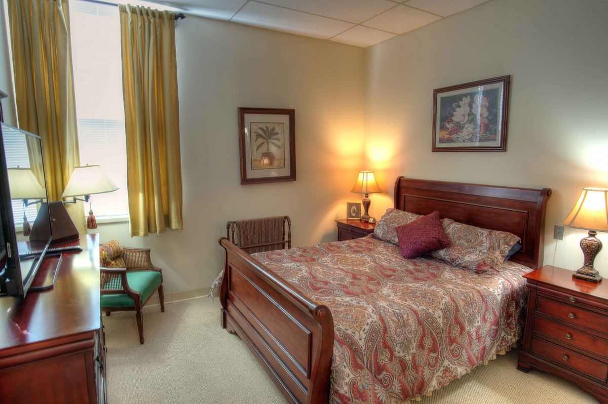 A cozy bedroom featuring a wooden bed, bedside lamp, artwork on the walls, and natural light filtering through curtains.