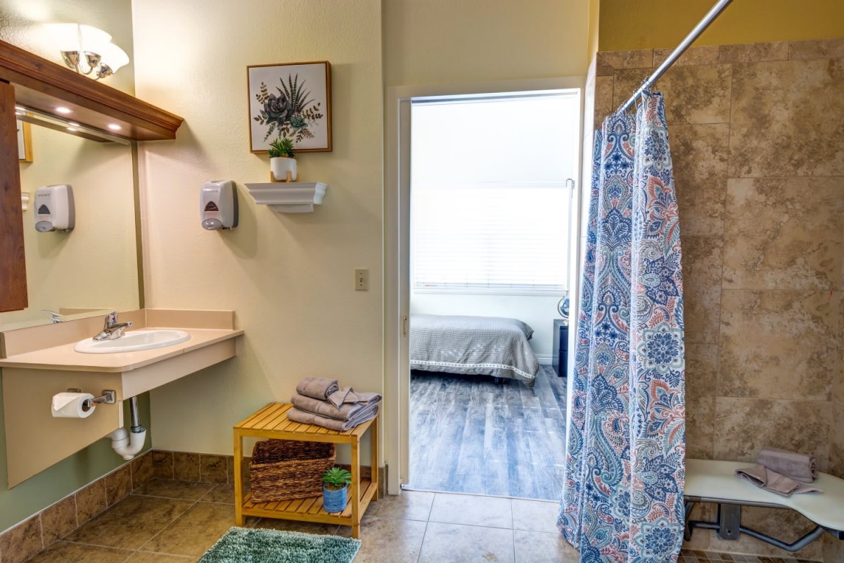 A view of a bathroom leading into a bedroom, featuring a shower curtain, sink, and neatly arranged towels.