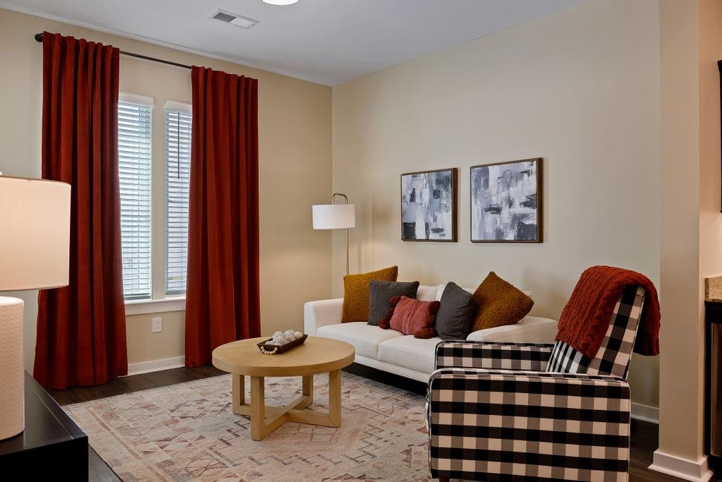 A cozy living room featuring a white sofa with colorful cushions, a round wooden coffee table, and a patterned armchair. Red curtains add a pop of color to the neutral walls, while two framed artworks hang above the sofa.