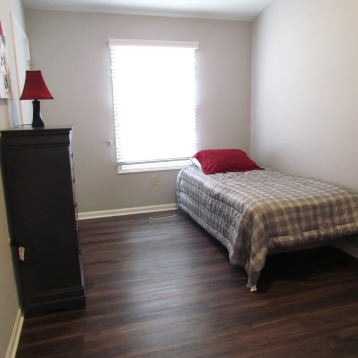 A minimalist bedroom with a single bed and dresser, featuring wooden floors and a window with natural light.