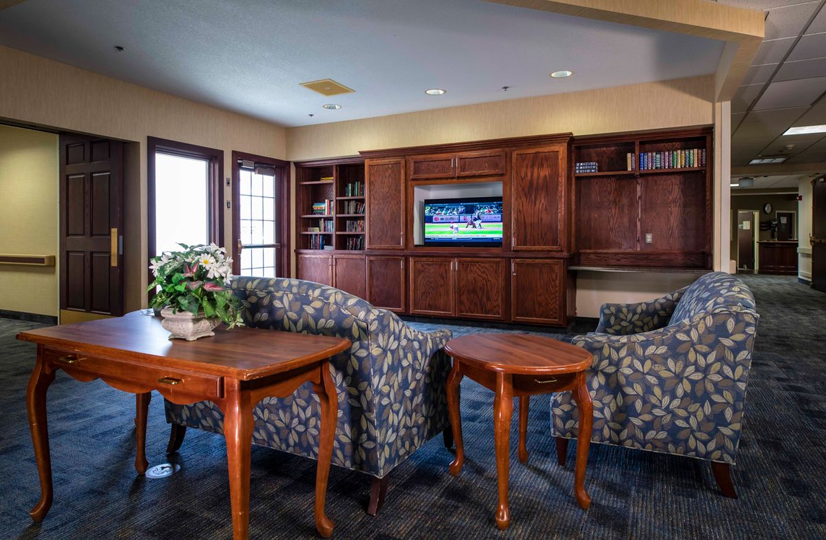 A cozy living room with comfortable chairs, wooden tables, and a television set against built-in bookshelves.