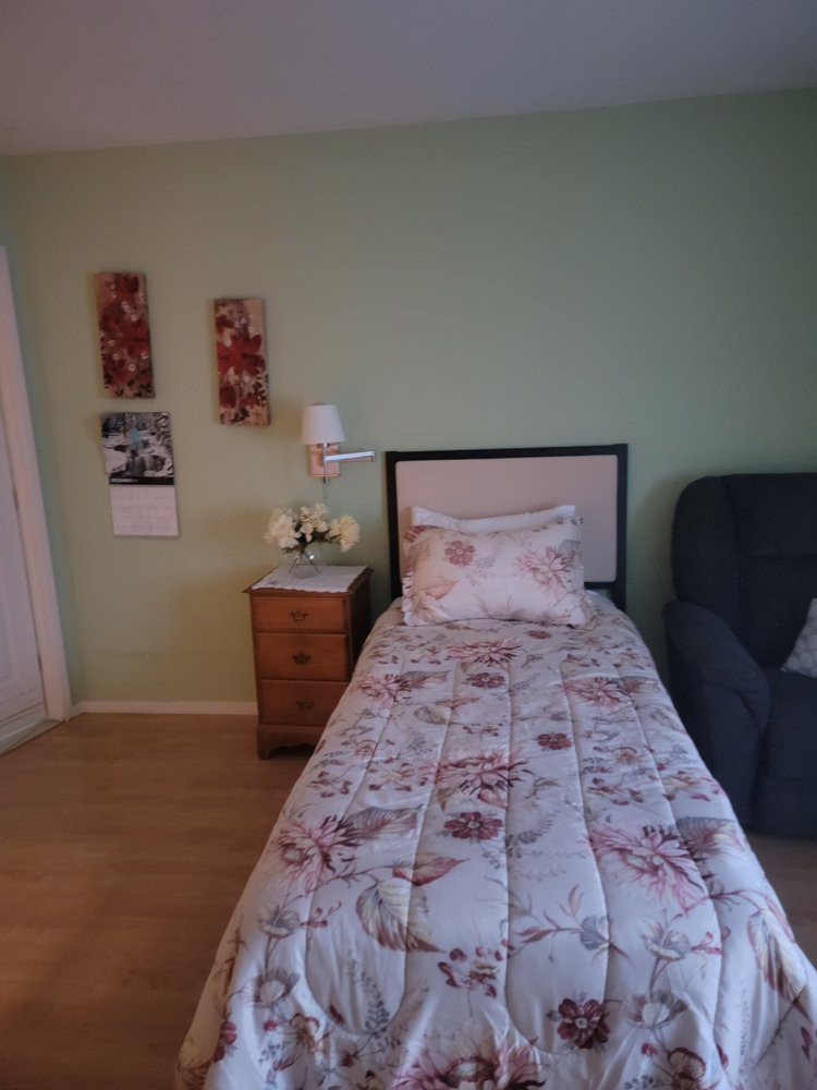 A cozy bedroom with a single bed featuring floral bedding, a nightstand with a lamp, and wall art.