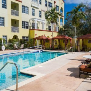 A sunny outdoor swimming pool area at a residential apartment building, surrounded by palm trees and lounge chairs.