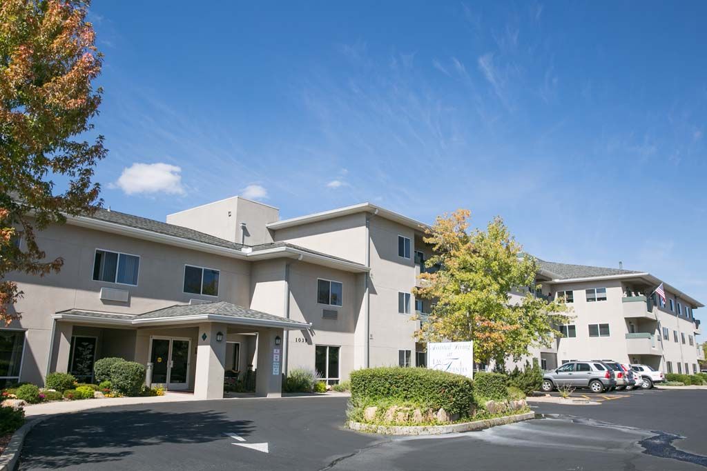 A multi-story building with an entrance, surrounded by trees and a parking area under a clear blue sky.