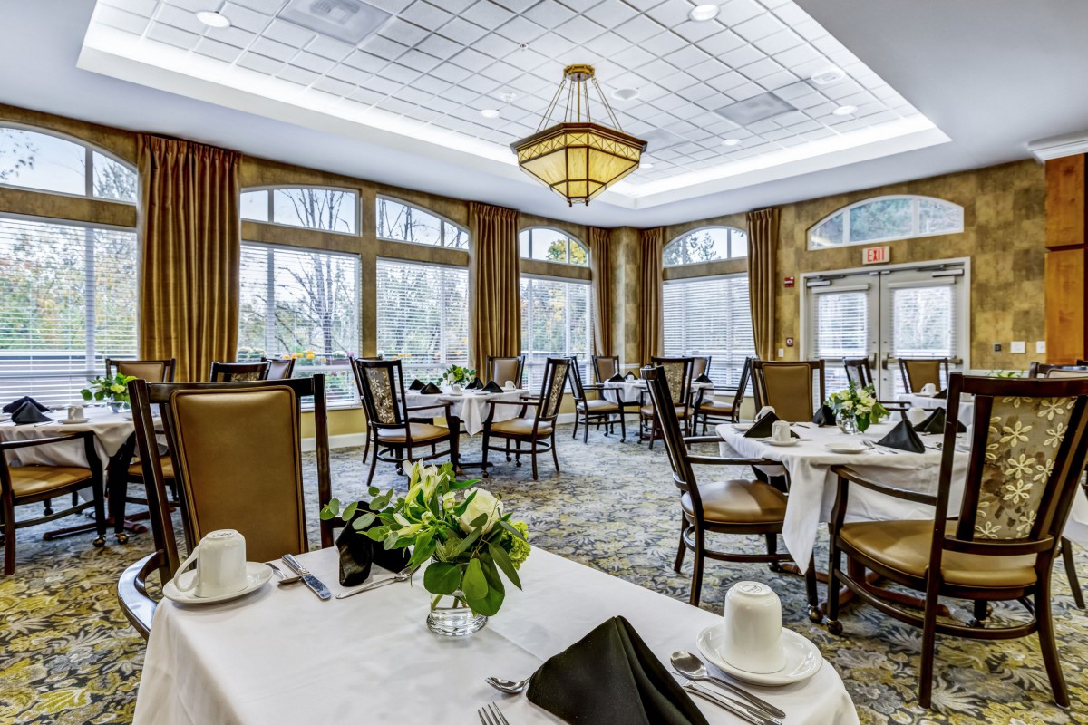 An elegant dining room with neatly arranged tables and chairs, featuring large windows and a chandelier.