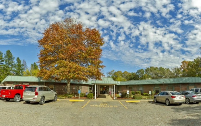 A small building with a parking lot and cars, surrounded by trees with autumn foliage under a partly cloudy sky.