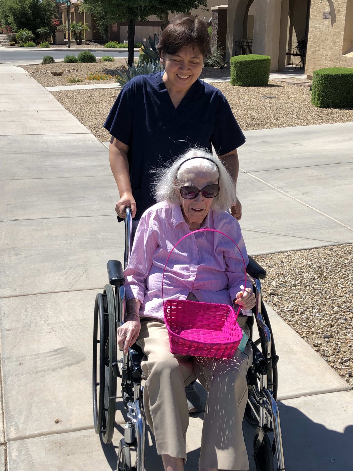An elderly woman smiling in a wheelchair, accompanied by a caregiver on a sunny day.