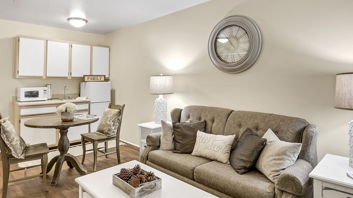 A cozy living room with a neutral color palette featuring a sofa, decorative pillows, and a large wall clock. A dining area is visible in the background.