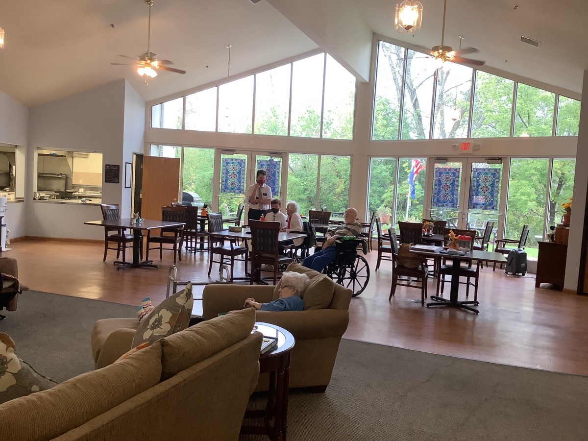 A spacious dining area with large windows, tables, chairs, and sofas, featuring ceiling fans and people seated, including one person in a wheelchair.