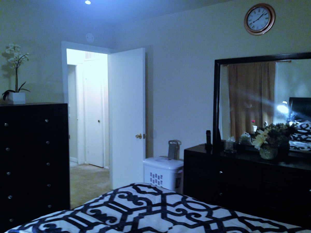 A bedroom featuring a bed with patterned bedding, a dresser with a mirror, and an open door leading to another room.