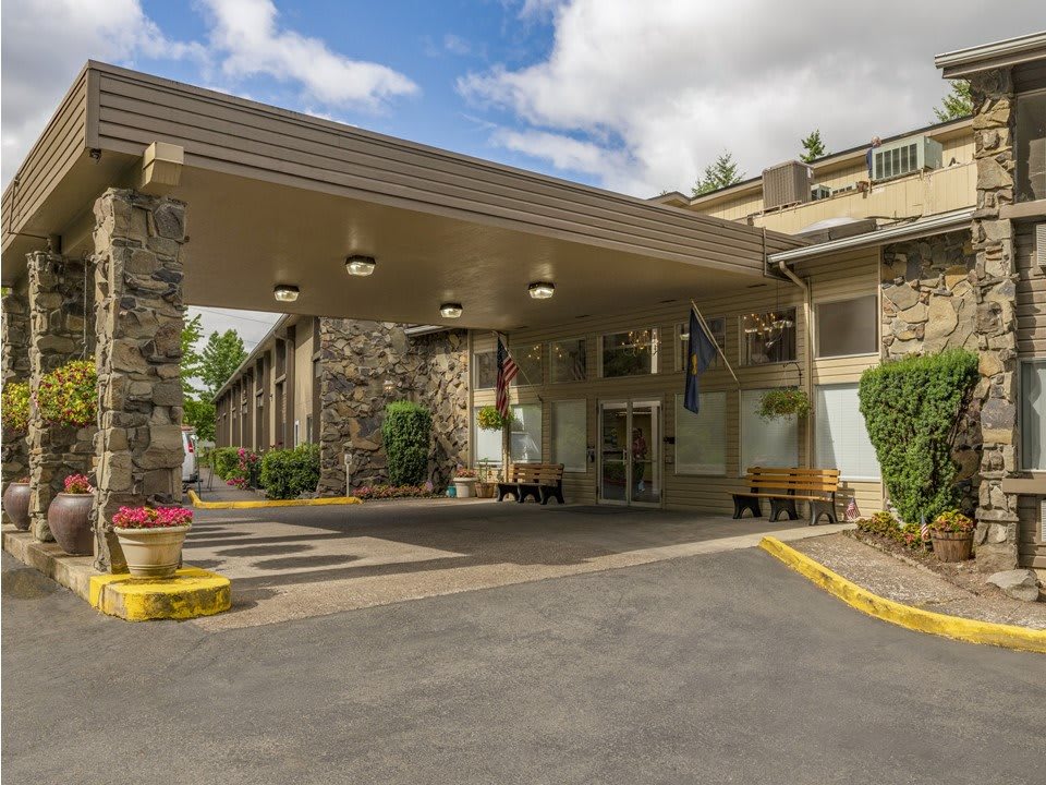 The entrance of a hotel with a wide canopy and stone accents, surrounded by landscaped greenery on a clear day.