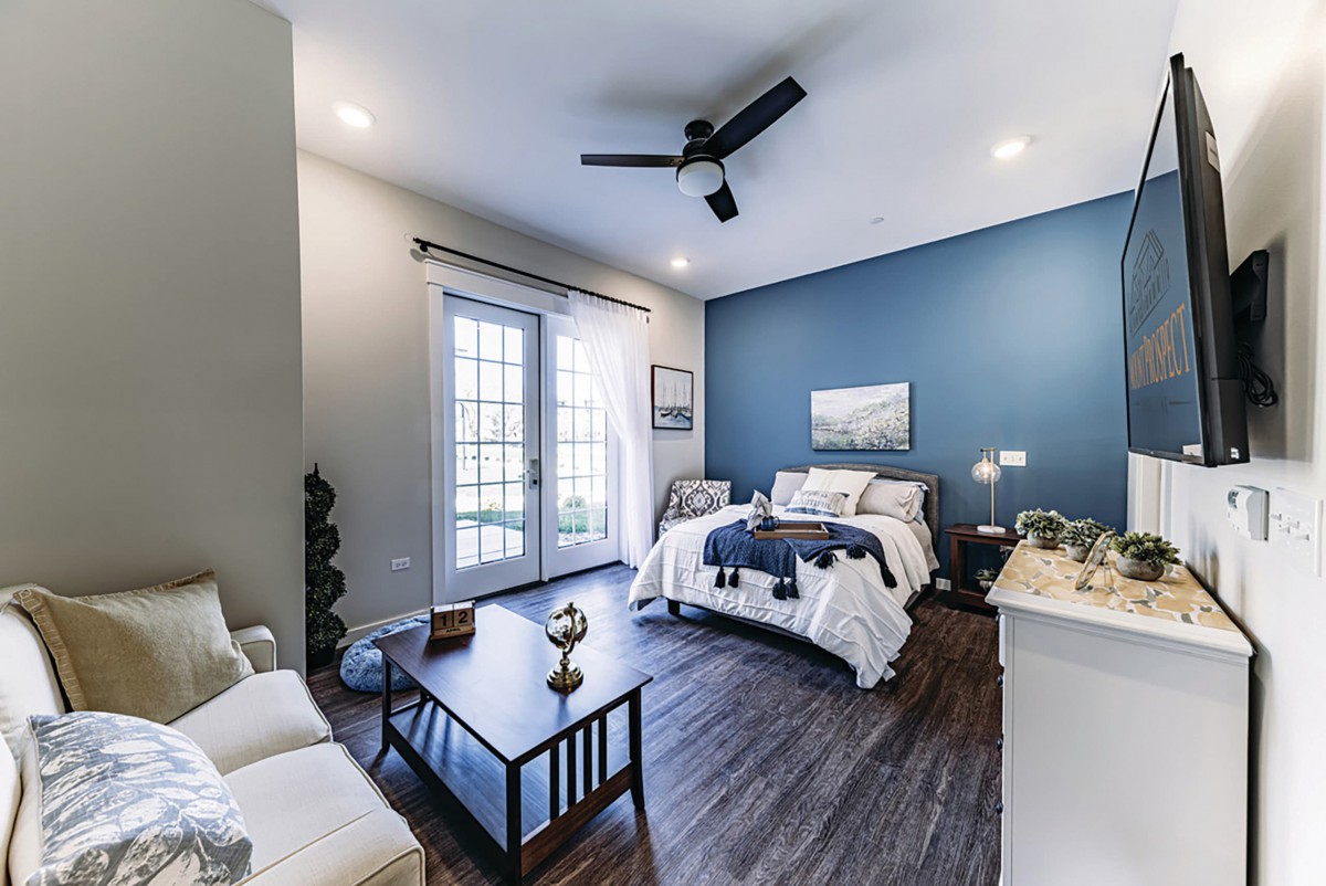 A modern bedroom with a blue accent wall, featuring wood flooring and abundant natural light through French doors.