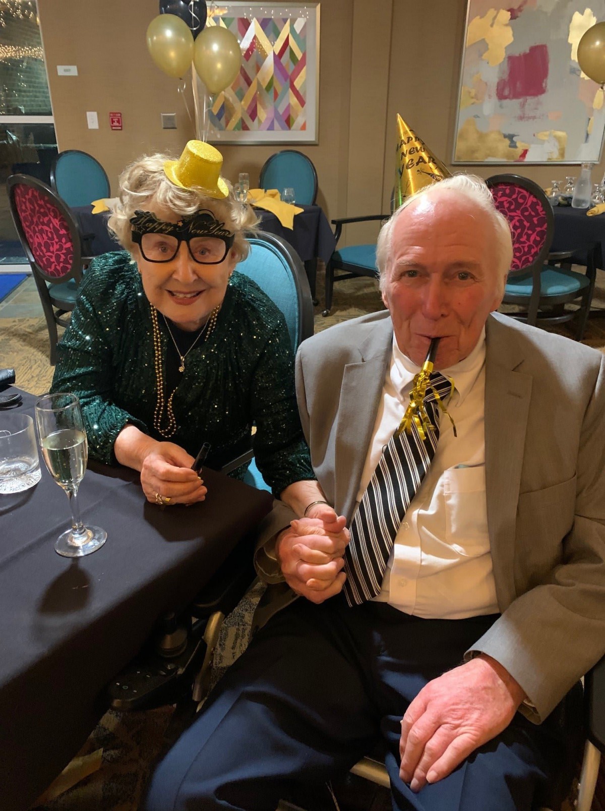 An elderly couple joyfully celebrating at a party, wearing festive hats and holding hands.