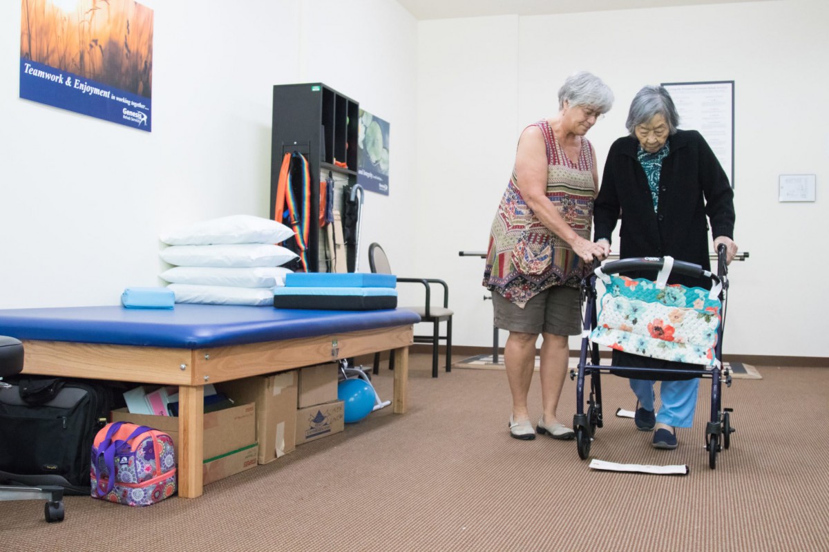 An elderly woman receives assistance from a caregiver while using a walker in a rehabilitation room.