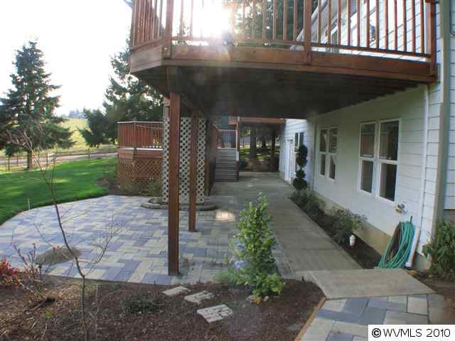 A backyard view featuring a patio area with a deck overhead, surrounded by greenery and landscaping.