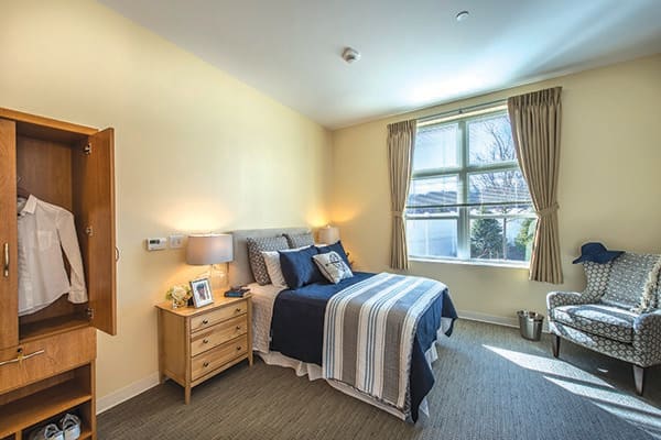 A cozy bedroom featuring a neatly made bed with striped bedding, a wooden nightstand with a lamp, and a large window with curtains letting in natural light.