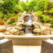 A tranquil garden scene featuring a small waterfall surrounded by lush greenery and rocks, viewed from a cozy patio.