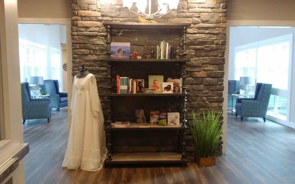 A cozy indoor setting featuring a bookshelf against a stone wall, with books, a dress mannequin, and a potted plant.