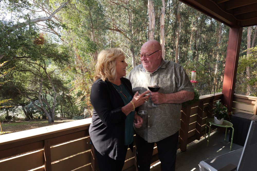 A couple enjoying a conversation on a balcony with wine and nature in the background.