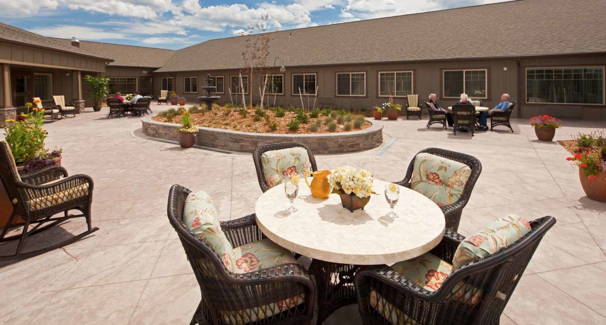 A serene courtyard with a round table and wicker chairs, surrounded by landscaping and a building in the background.