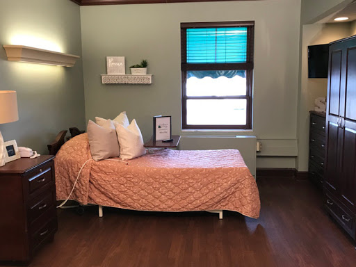 A cozy hospital room featuring a single bed with an orange quilt and a window offering natural light.