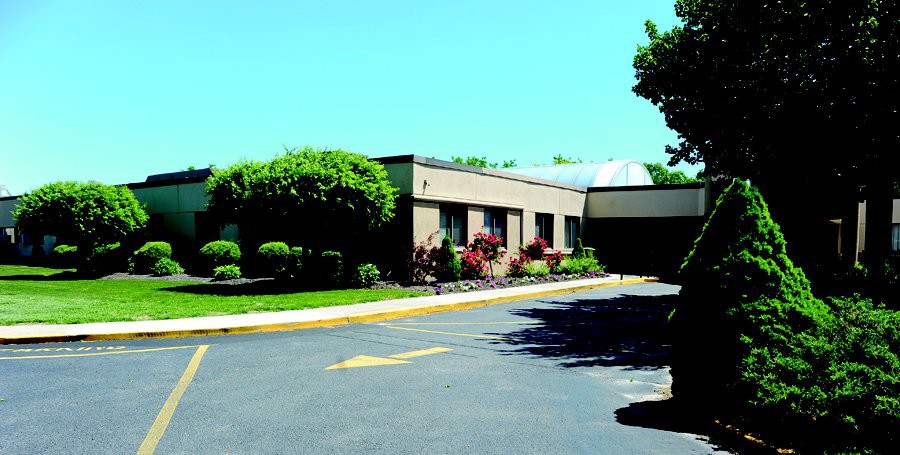 A commercial building surrounded by trees and bushes, with a road in the foreground.
