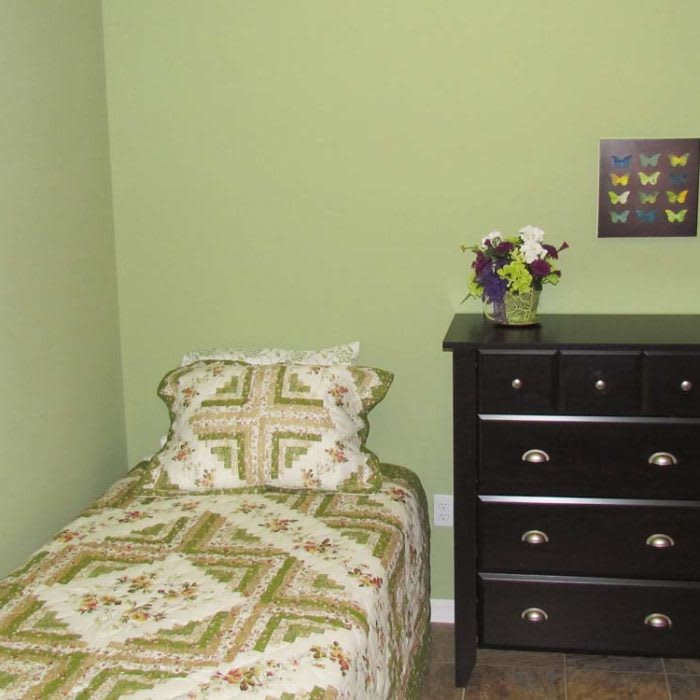 A cozy bedroom with green walls, featuring a single bed with a patterned quilt and a black dresser topped with flowers.