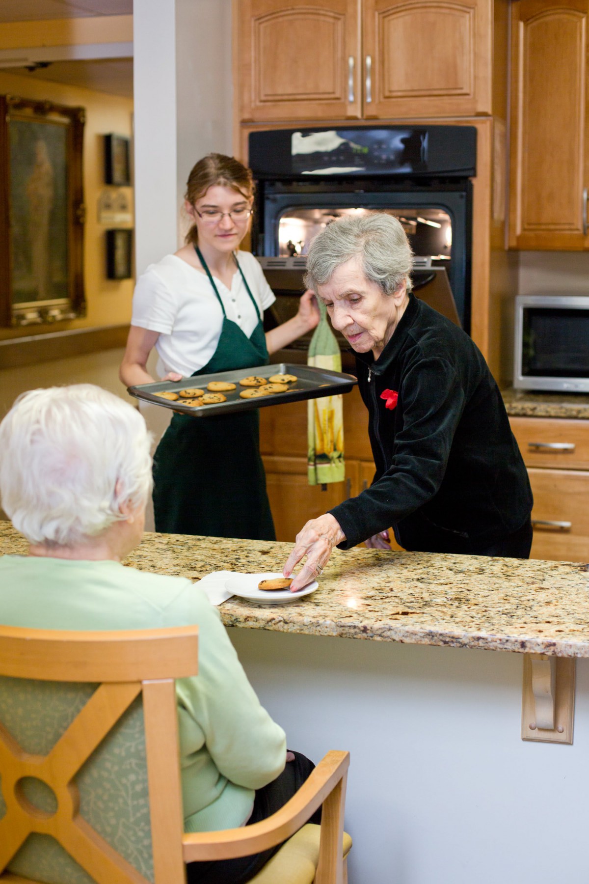 Two women baking cookies in a kitchen, with one serving an elderly person.