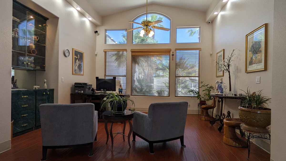 A cozy living room with large windows, featuring comfortable chairs, plants, and a ceiling fan.