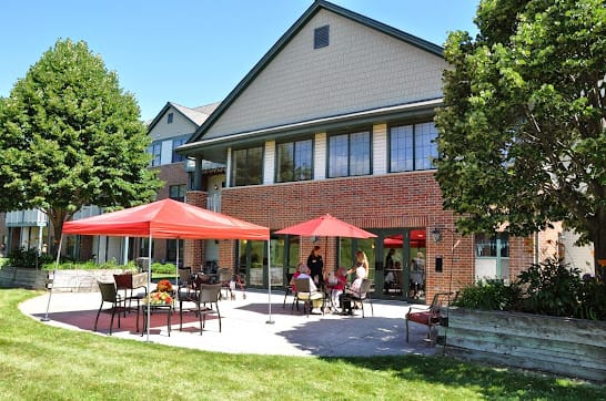 A sunny patio area with red umbrellas and people enjoying the outdoor seating in front of a brick building.