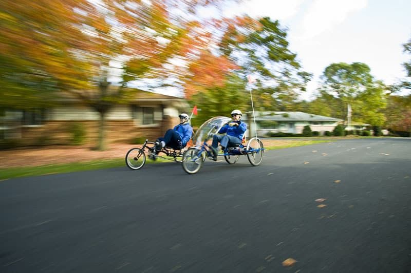 Two people riding recumbent bicycles down a tree-lined street in autumn.