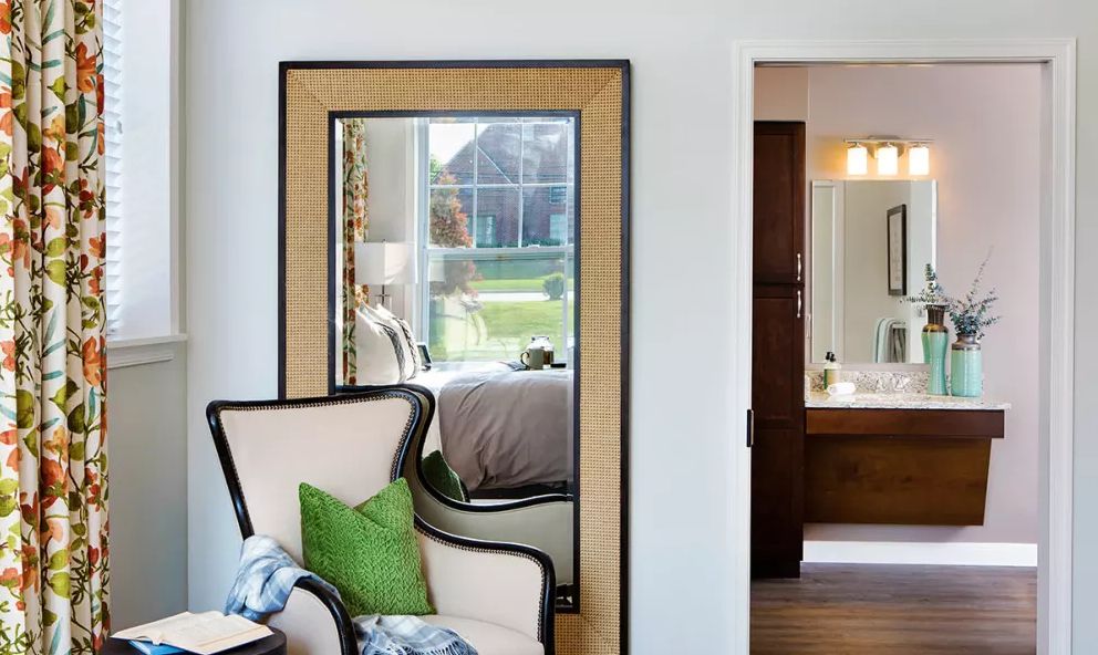 A cozy bedroom featuring a large framed mirror reflecting the room's decor, with a stylish chair and green pillow in the foreground.