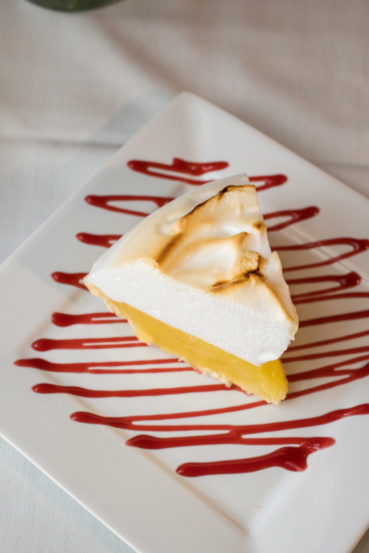 A slice of lemon meringue pie drizzled with raspberry sauce on a plate.