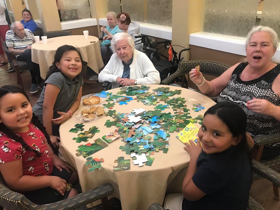 A group of people, including children and an elderly person, enjoy working on a puzzle together at a round table.