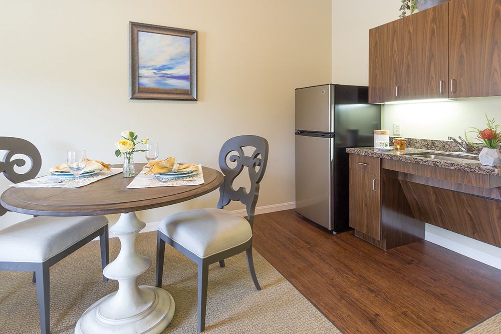 A cozy dining area with a round table, two chairs, and a compact kitchen setup featuring a refrigerator and wooden cabinets.