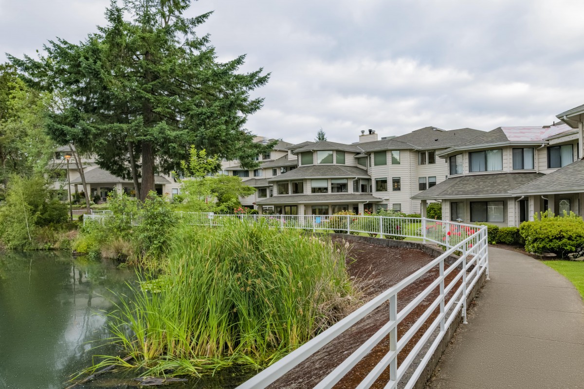 A scenic view of residential buildings surrounded by greenery and a pond, with a walkway leading through the area under a cloudy sky.