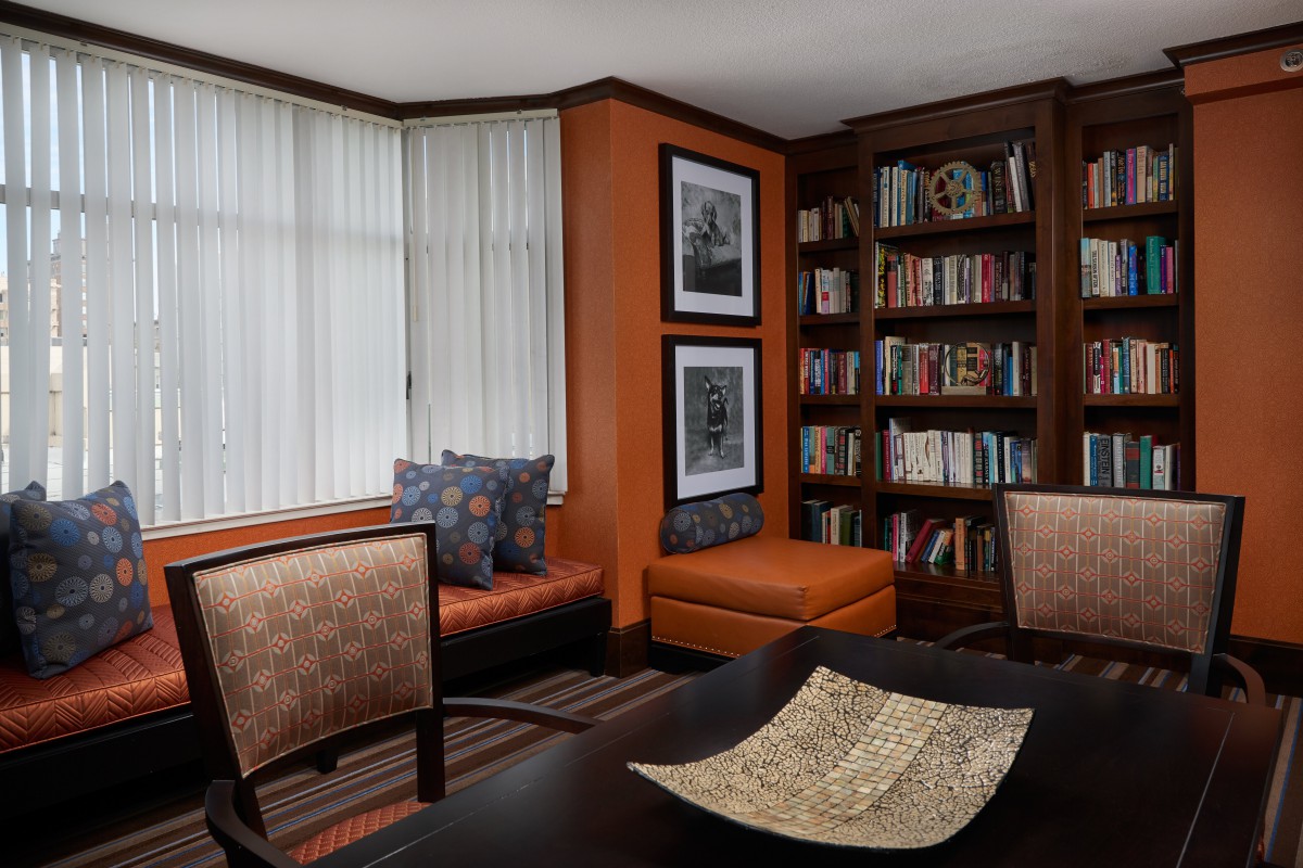 A cozy office space with orange walls, featuring a bookshelf, window seat, table, and chairs.