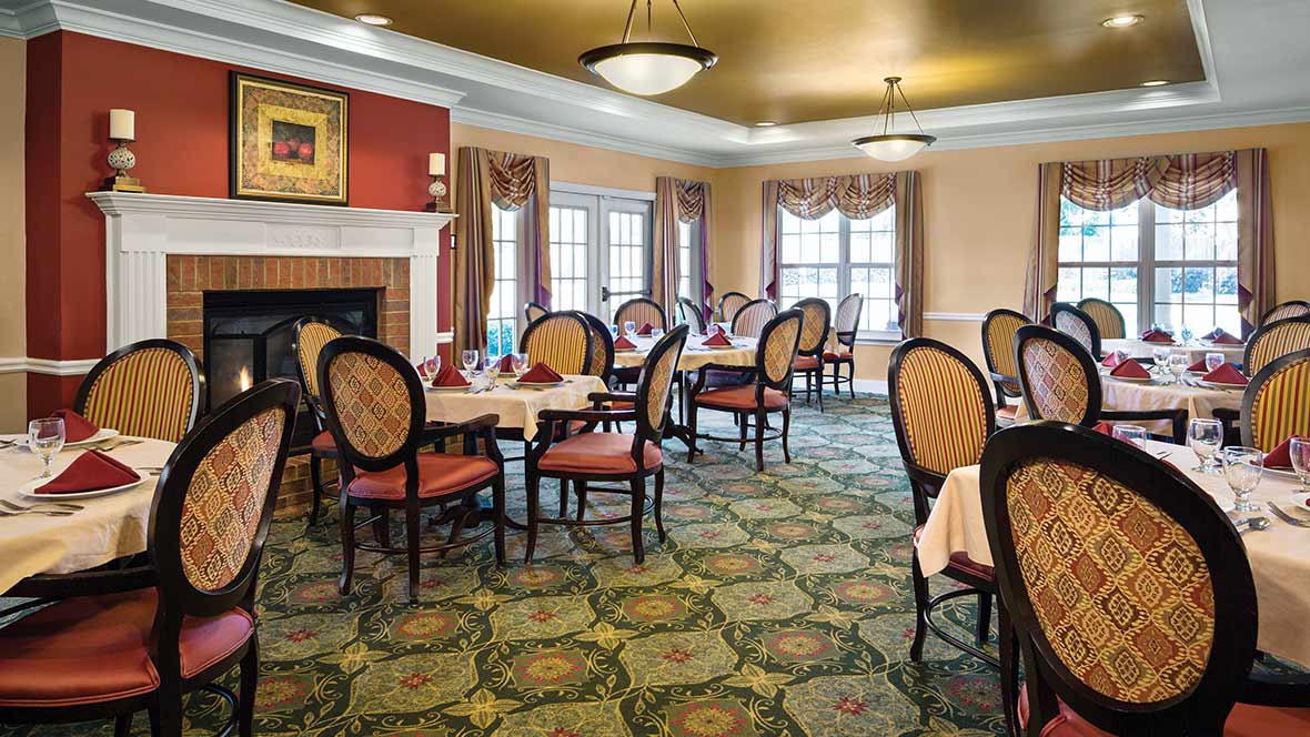 An elegant dining room with neatly arranged tables and chairs, featuring large windows with curtains and a decorative carpet.