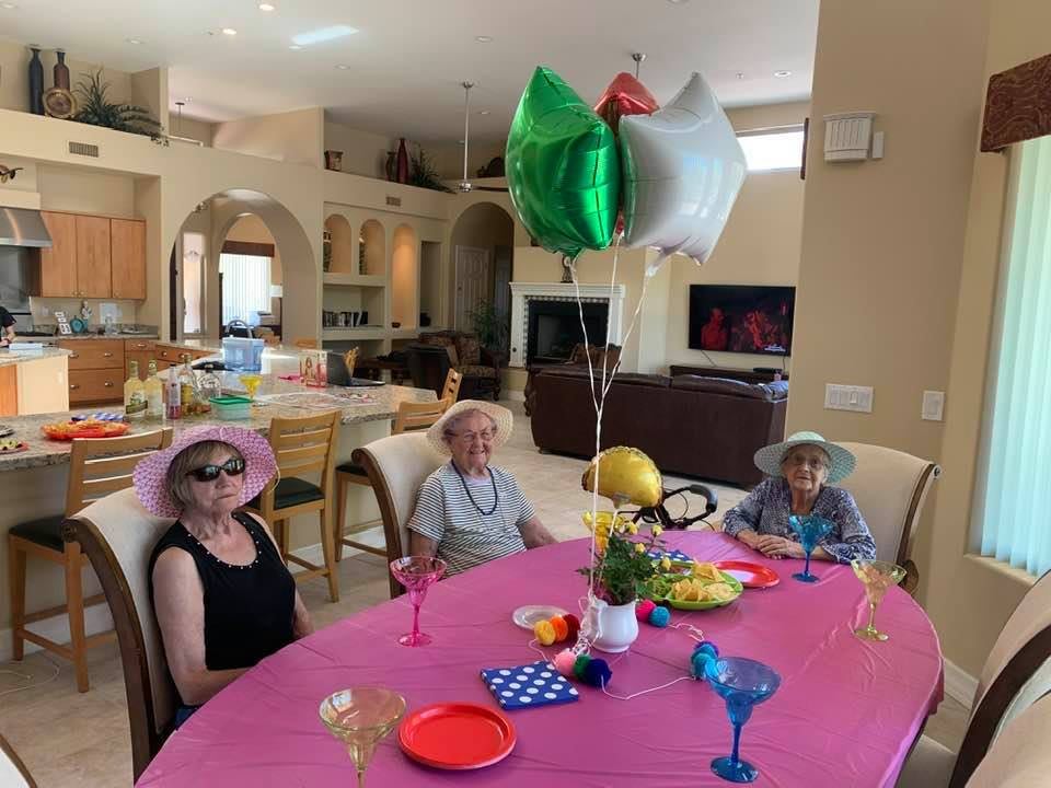 A festive indoor gathering with people sitting around a decorated table, adorned with balloons and colorful decorations.