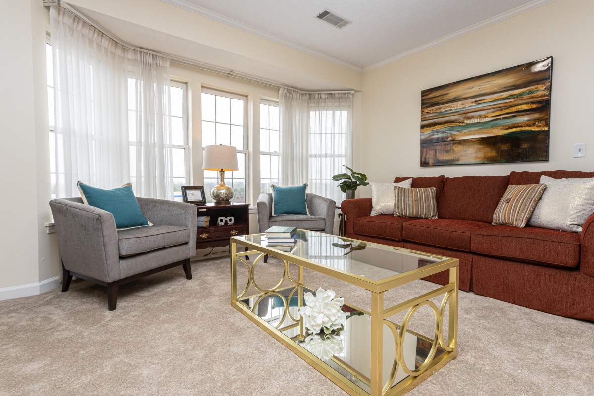 A cozy living room with a brown sofa, two gray armchairs, and a glass coffee table adorned with decor. Large windows bring in natural light, and a piece of artwork adds color to the walls.