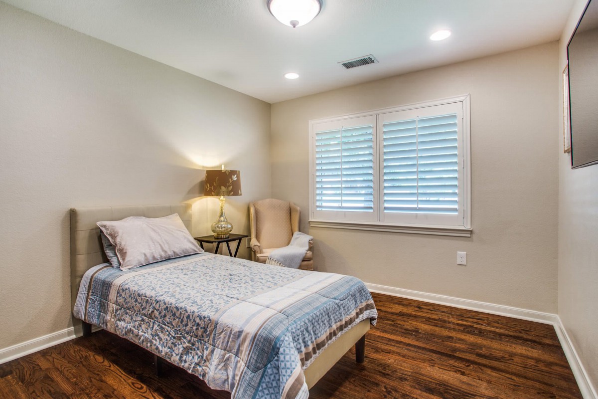 A cozy bedroom with a single bed, bedside lamp, chair, and a window with shutters.