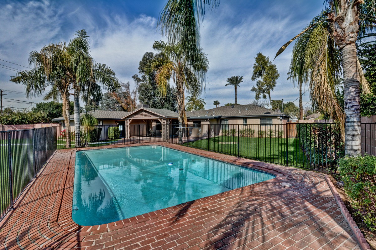 A backyard oasis featuring a swimming pool surrounded by palm trees and a brick patio.