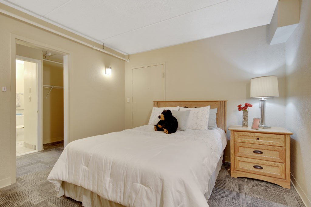 A cozy bedroom with a neatly made bed, featuring a small dog sitting on it, next to a wooden nightstand with a lamp and flowers.
