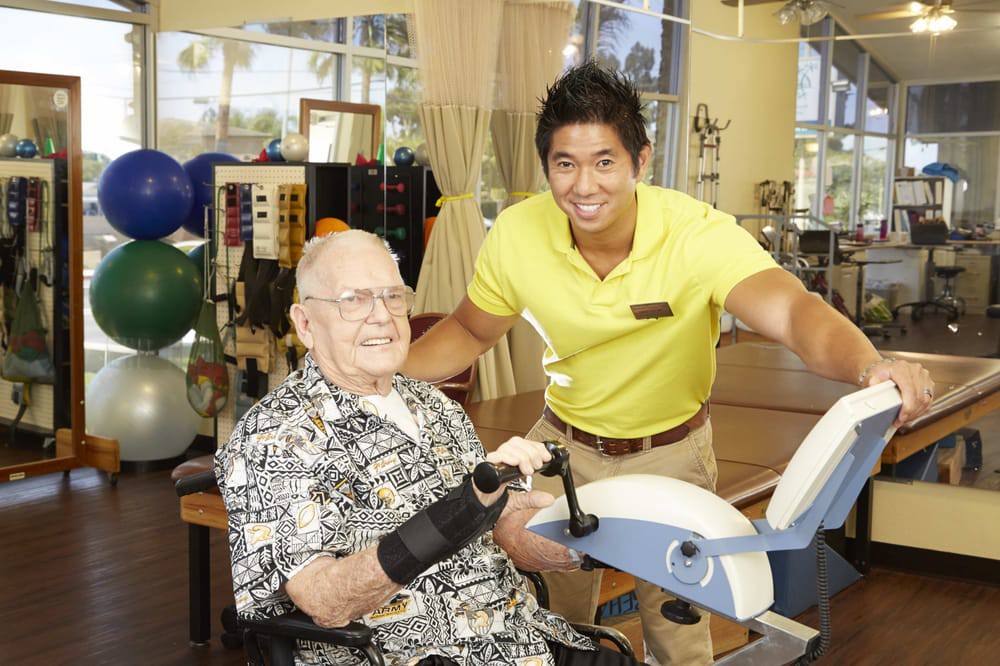 A healthcare professional assists an elderly person during a physical therapy session with exercise equipment.